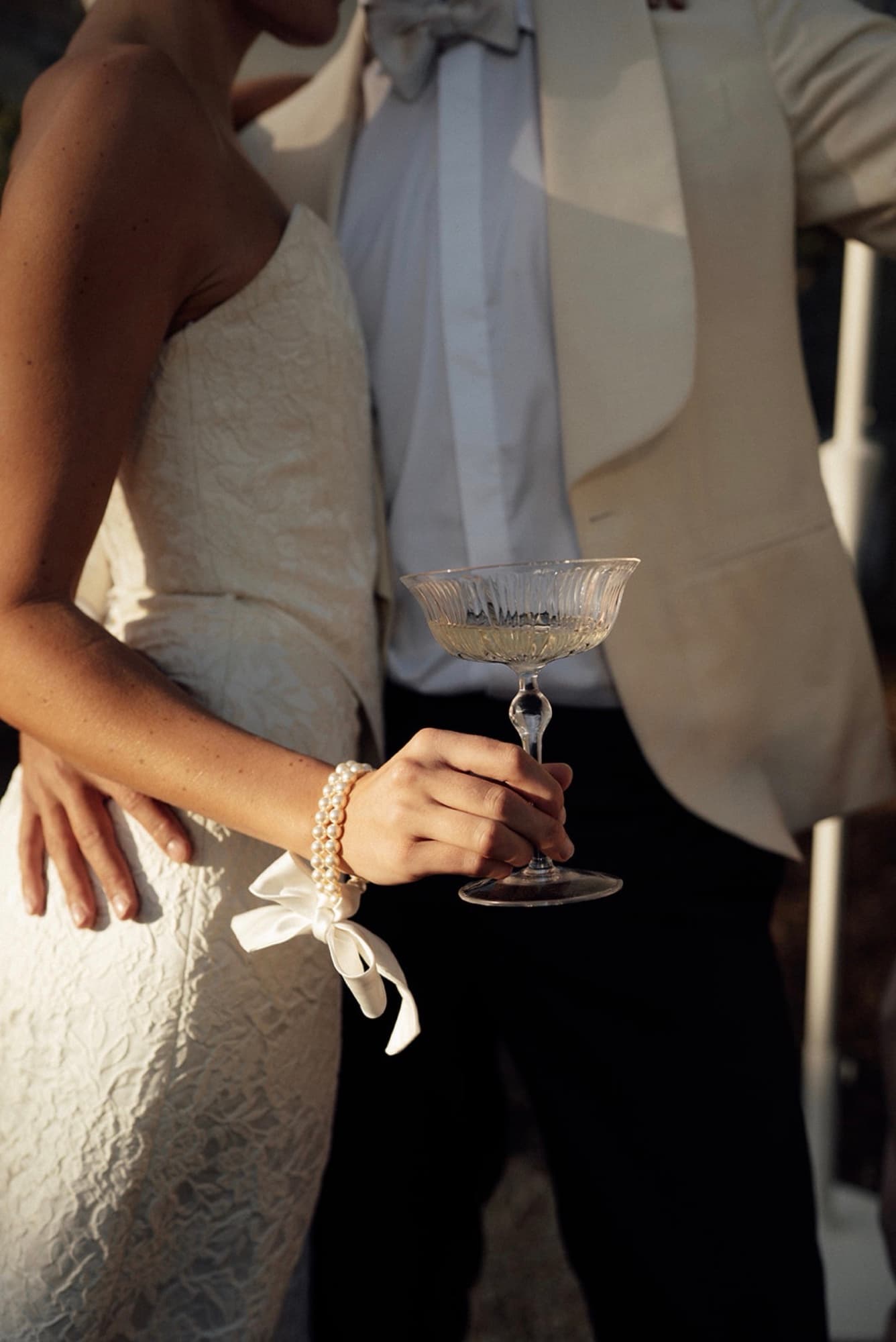 A champagne coupe in hand at golden hour — brocade gown, pearls and a white dinner jacket
